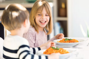 Smiling Children Having Fun While Eating Spaghetti With Tomato Sauce In The Kitchen At Home.