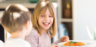 Smiling Children Having Fun While Eating Spaghetti With Tomato Sauce In The Kitchen At Home.