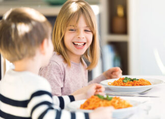 Smiling Children Having Fun While Eating Spaghetti With Tomato Sauce In The Kitchen At Home.