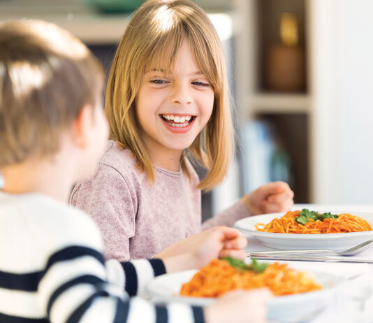 Smiling Children Having Fun While Eating Spaghetti With Tomato Sauce In The Kitchen At Home.