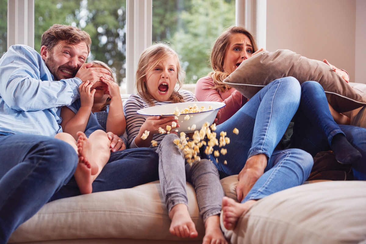 Frightened Family Sitting On Sofa At Home Watching Horror Movie On Tv With Popcorn