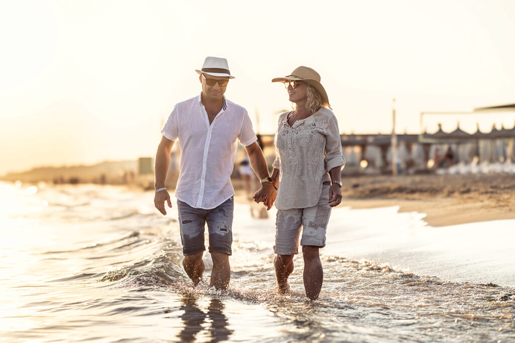 A Middle Aged Couple Walking Hand In Hand On The Beach At Sunset.