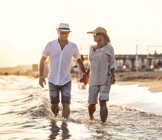 A Middle Aged Couple Walking Hand In Hand On The Beach At Sunset.