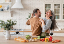 Happy Cheerful Middle Aged Mature Couple Family Parents Dancing Together In The Kitchen, Preparing Cooking Food Meal For Romantic Dinner, Spending Time Together. Active Seniors