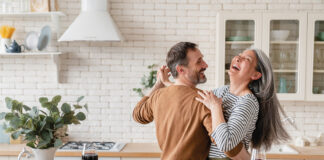 Happy Cheerful Middle Aged Mature Couple Family Parents Dancing Together In The Kitchen, Preparing Cooking Food Meal For Romantic Dinner, Spending Time Together. Active Seniors