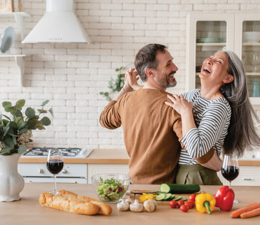 Happy Cheerful Middle Aged Mature Couple Family Parents Dancing Together In The Kitchen, Preparing Cooking Food Meal For Romantic Dinner, Spending Time Together. Active Seniors